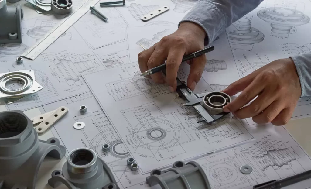 Engineers hands measuring a bearing with caliper over detailed mechanical blueprints and parts overviewing the workspace